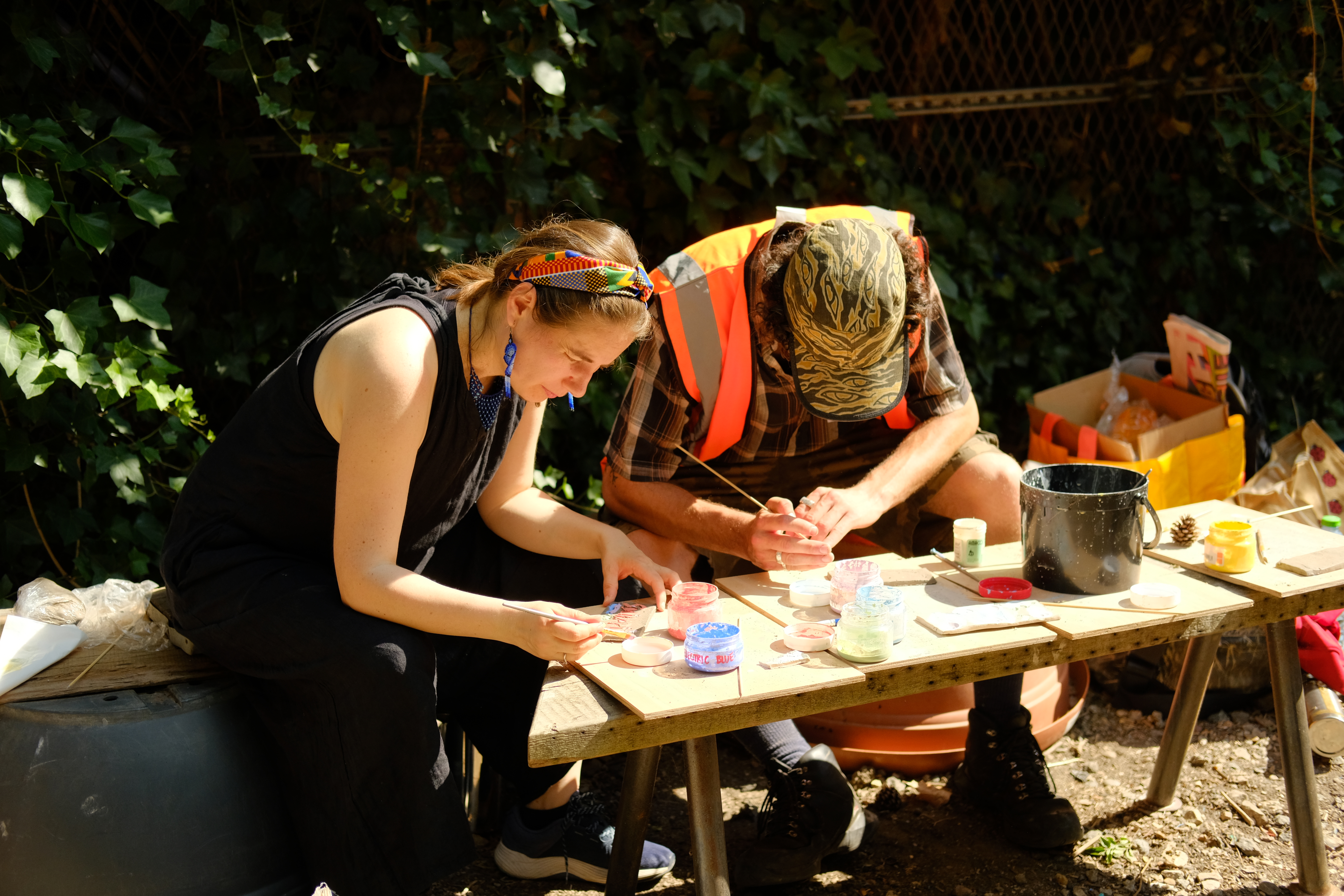 Two people sit outdoors at a small table, working together on a hands‑on craft activity using paints and various small materials